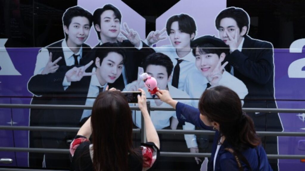 BTS fans take photos of BTS merchandise with a BTS banner displayed on the HYBE building in the background