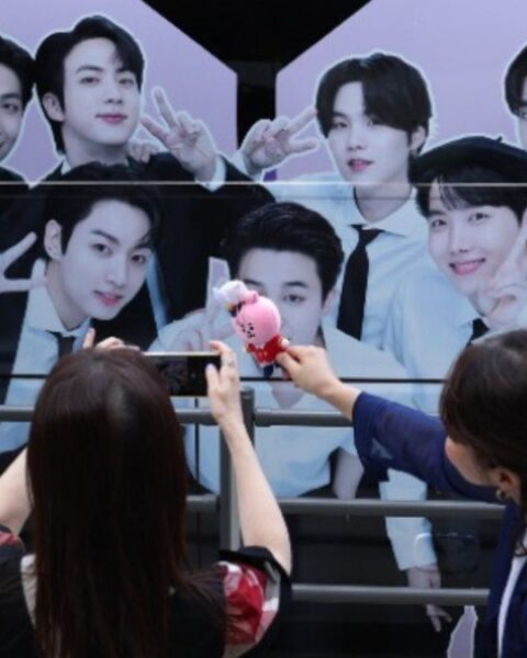 BTS fans take photos of BTS merchandise with a BTS banner displayed on the HYBE building in the background